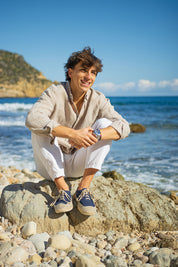 Modelo frente al mar sentado en las rocas con las alpargatas Peñiscola Azul de MYBARKS, unas alpargatas estilo sneaker de caña baja para hombre hechas con materiales sostenibles perfectas para el verano. Yute y lino natural con aroma a cítricos mediterráneos. 